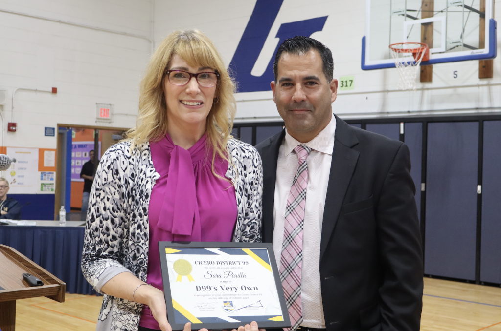 A smiling woman and man hold a certificate together in a school gymnasium.